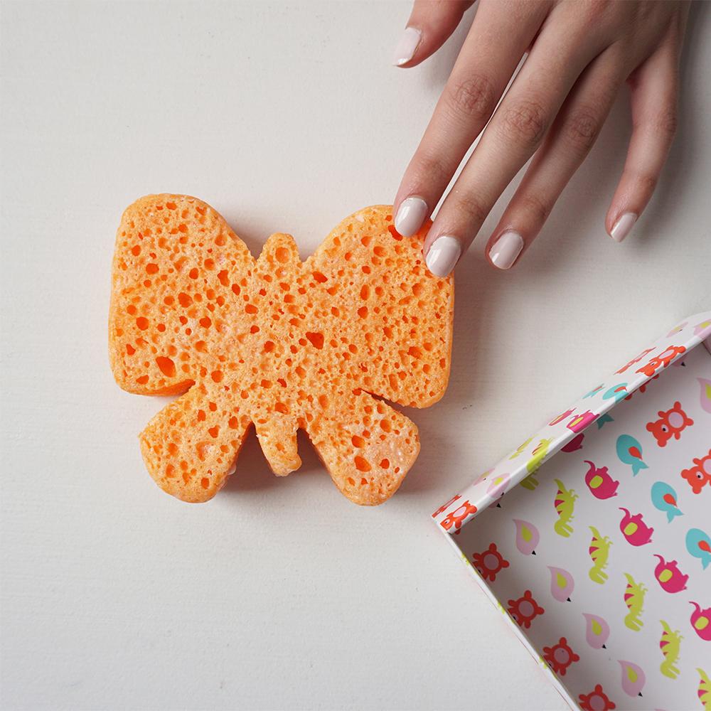 Een hand raakt de oranje badspons in vlindervorm aan, naast een doos met kleurrijke dierenpatronen.