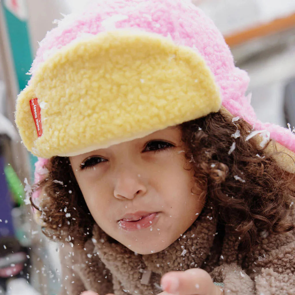 Close-up van een kind met de roze en gele Cozy Kira muts dat sneeuw wegblaast.