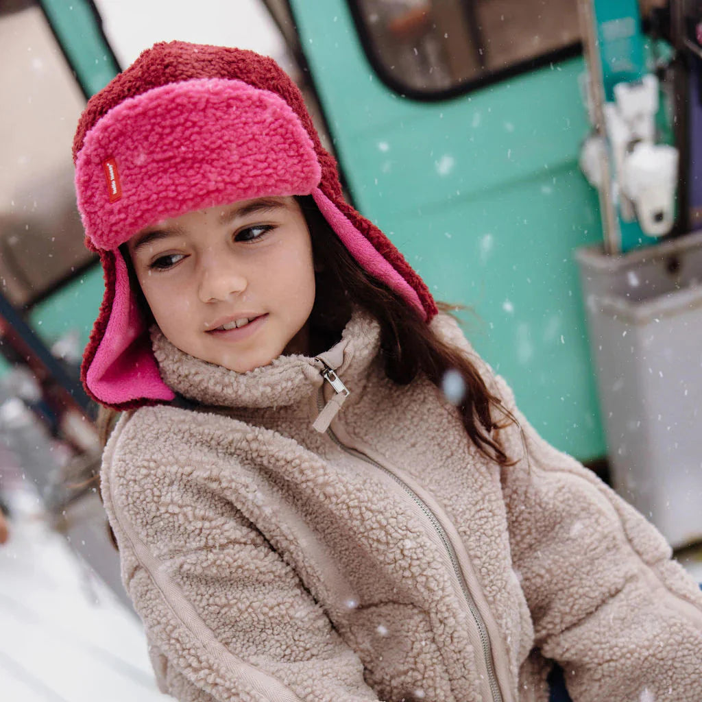 Kind draagt een bordeauxrode en roze Cozy Tara chapka met oorkleppen en een beige jas in de sneeuw.