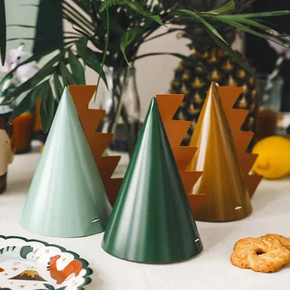 Drie dino-feesthoedjes in lichtgroen, donkergroen en okergeel met oranje stekels op een feesttafel.