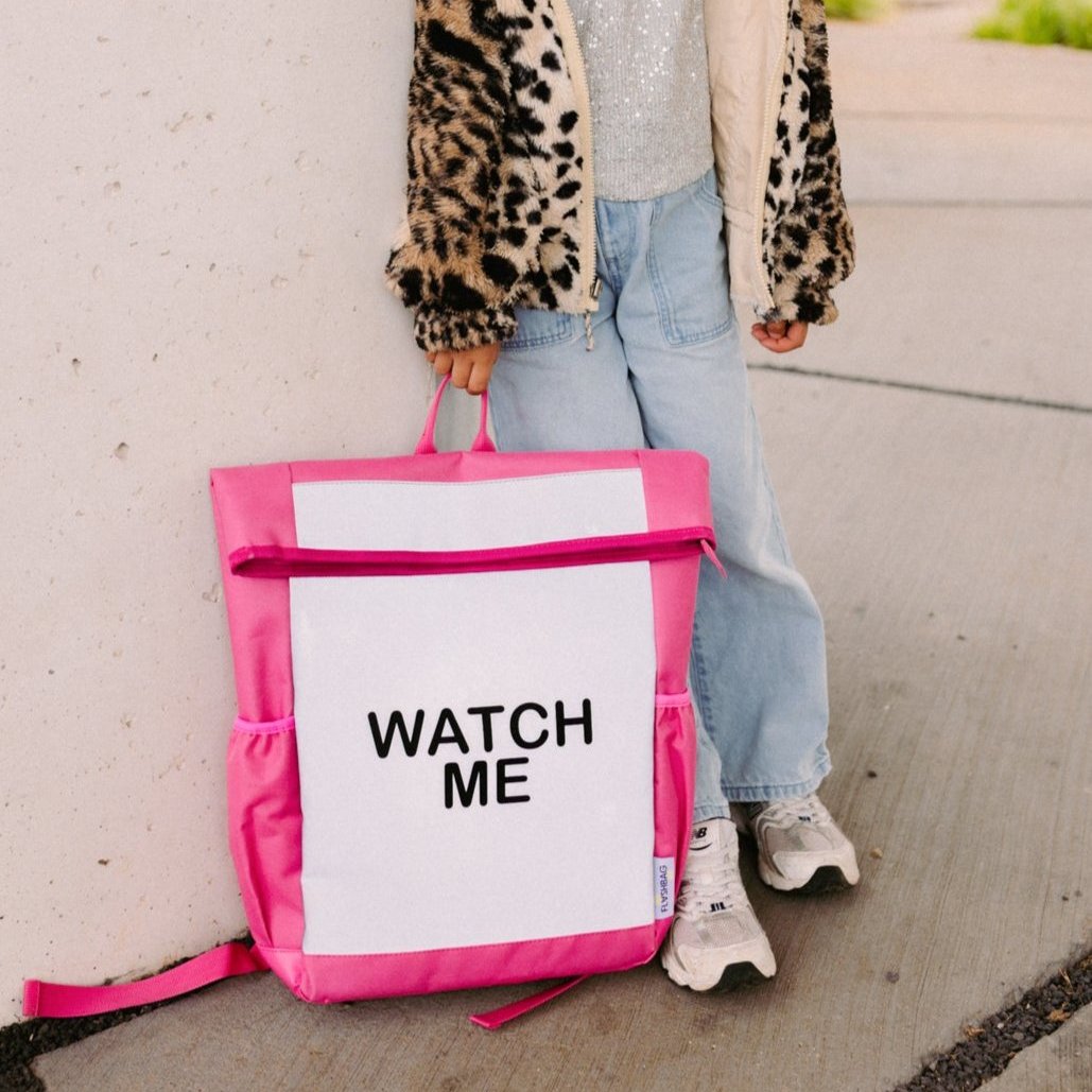 Roze Flashbag Lou-rugzak met grijs voorpaneel, vastgehouden aan het handvat door een kind naast een muur.