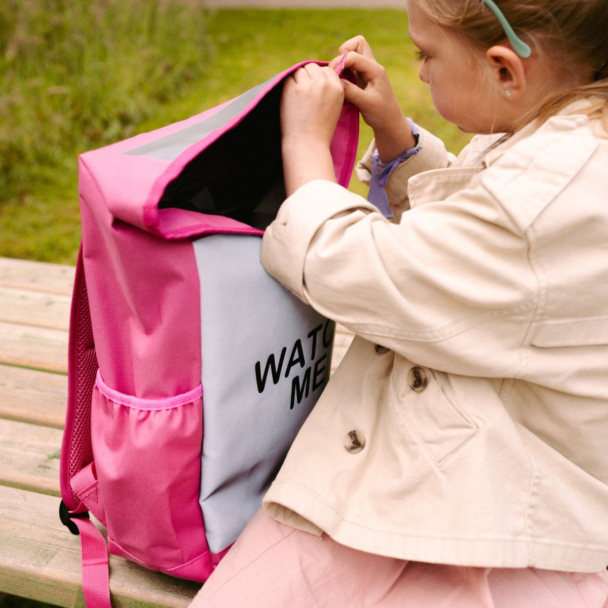 Kind opent de roze Flashbag Lou-rugzak op een houten bank, met zicht op de zwarte binnenkant en het zijvak.