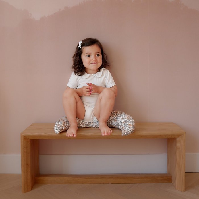 Kind met een wit haarspeldje met strik, zittend op een houten bank tegen een roze muur.