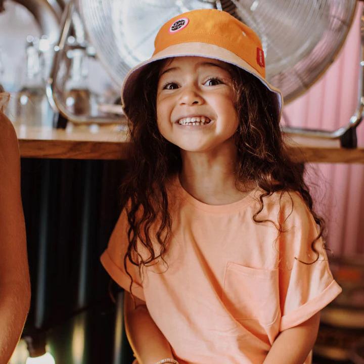 Lachend kind met het oranje hoedje Mini Orange van Hello Hossy en een perzikkleurig T-shirt.