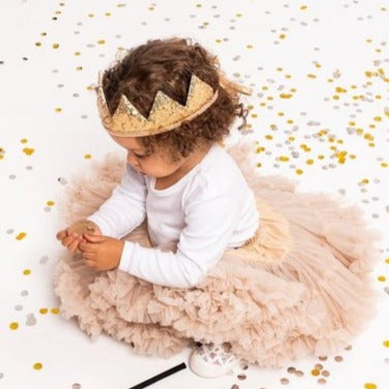 Kind met een gouden paillettenkroon, een witte top en een lichtroze rok tussen gouden confetti.