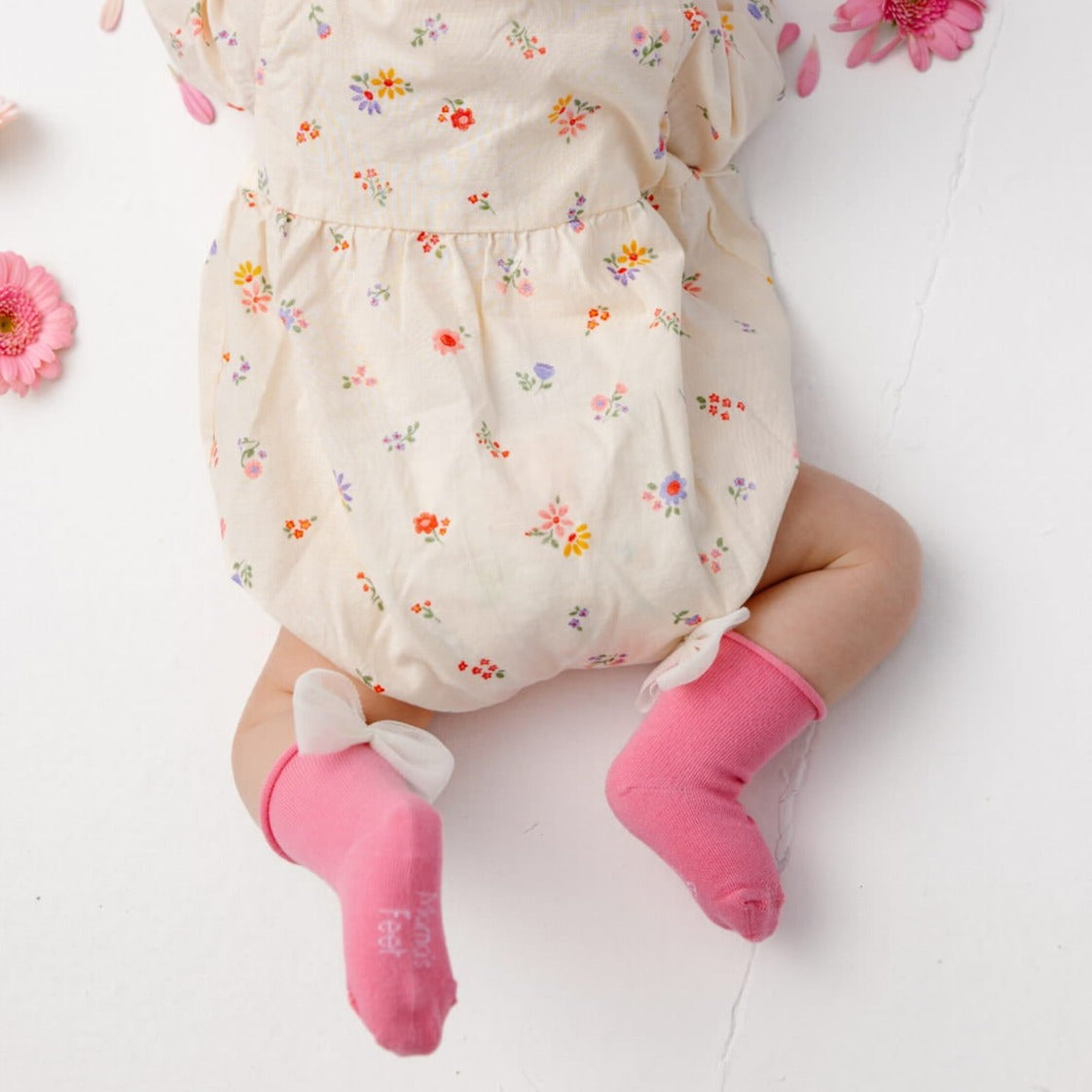 Baby in een crèmekleurige outfit met bloemenprint en roze Magnolia sokken met witte strikken.