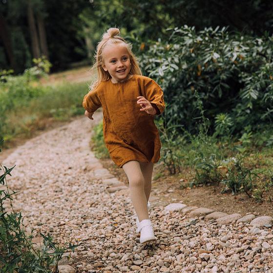 Kind met beige geribde maillot, okerkleurige jurk en witte schoenen op een bospad.