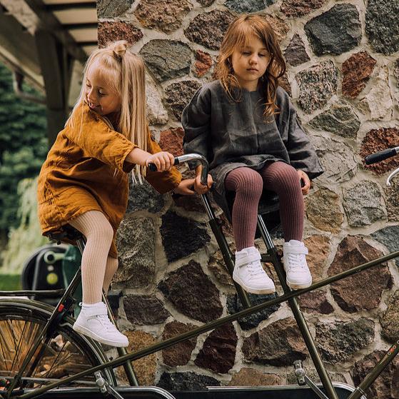 Twee kinderen op een fiets bij een stenen muur, één met beige geribde maillot en één met een paarse maillot.