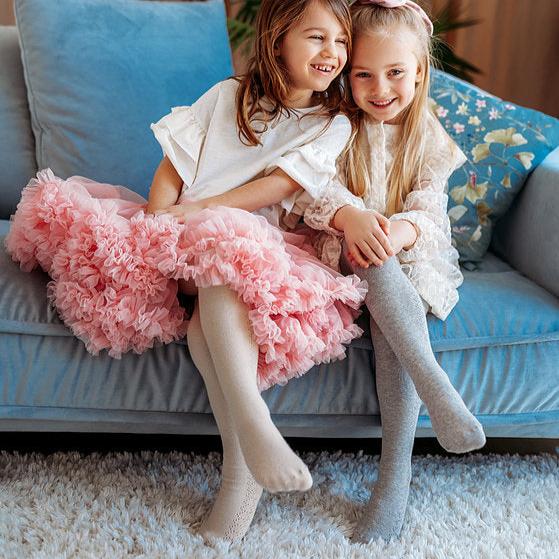 Twee meisjes op een blauwe bank dragen de maillot Josephine in beige en grijs onder een roze rok en witte jurk.