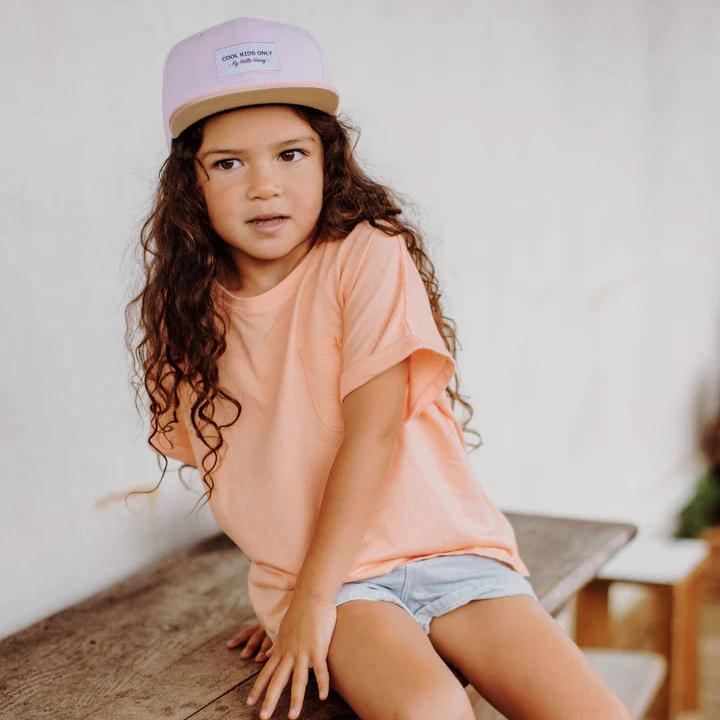 Kind met Hello Hossy Mini Purple pet, perzikkleurig shirt en korte broek, zittend op een houten bank.
