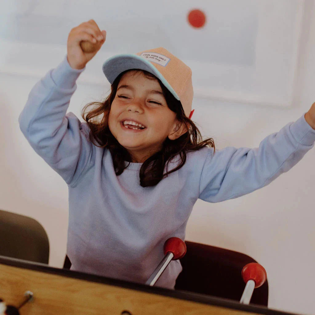 Kind met de perzikkleurige Sweet Peach pet en mintgroene klep, met armen omhoog bij een tafelvoetbalspel.
