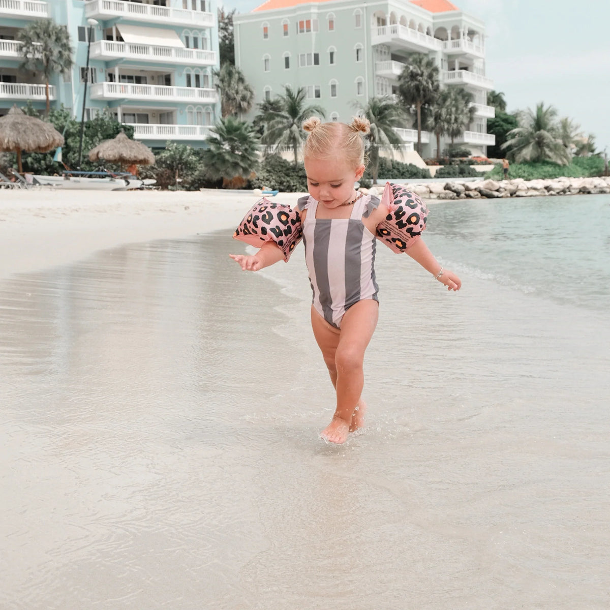 Kind met rosékleurige zwembandjes met panterprint loopt door ondiep zeewater bij het strand.