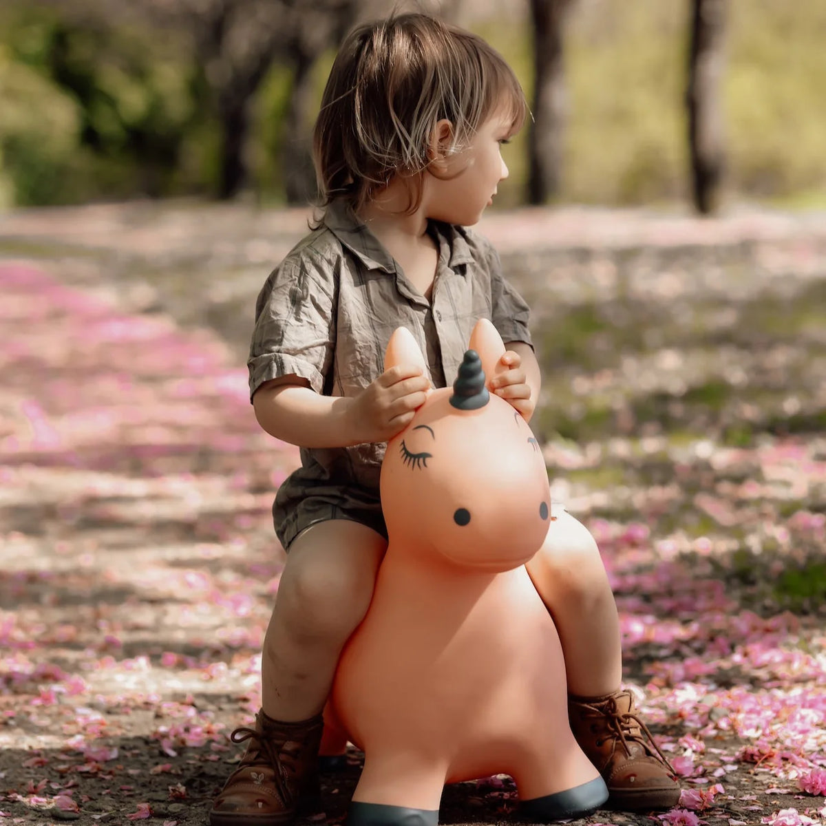 Kind zit buiten op het zachtroze springdier Luna de eenhoorn met antracietgrijze hoorn, op een pad met roze bloesem.