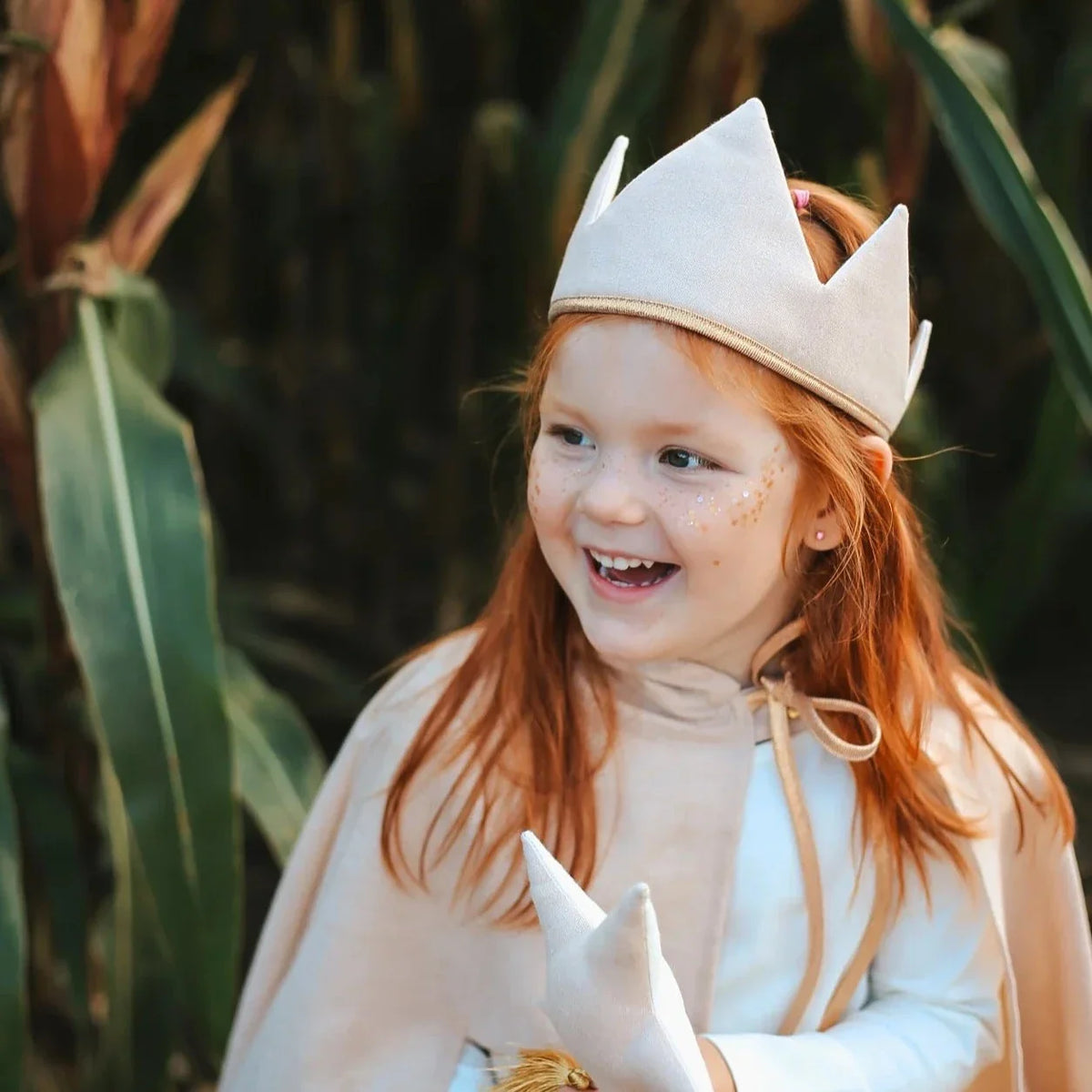 Kind met gouden gezichtsglitter, een lichte kroon en cape tussen hoge groene planten.