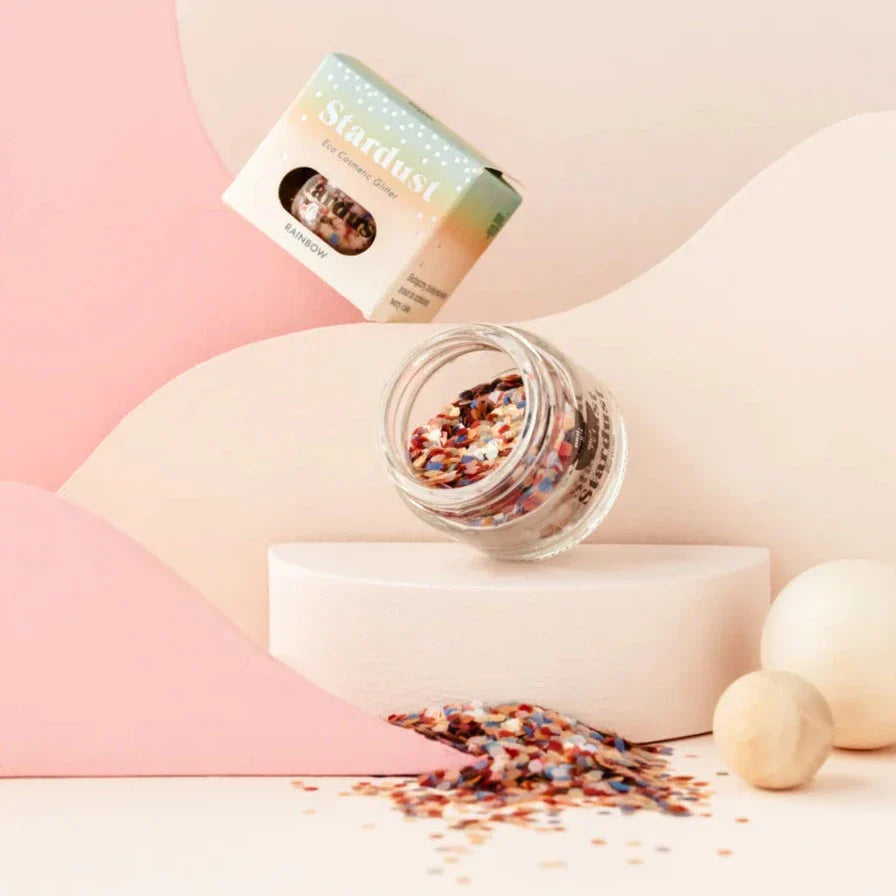Open glazen pot Star Dust Rainbow gezichtsglitter met verpakking en uitgestrooide veelkleurige glitters.