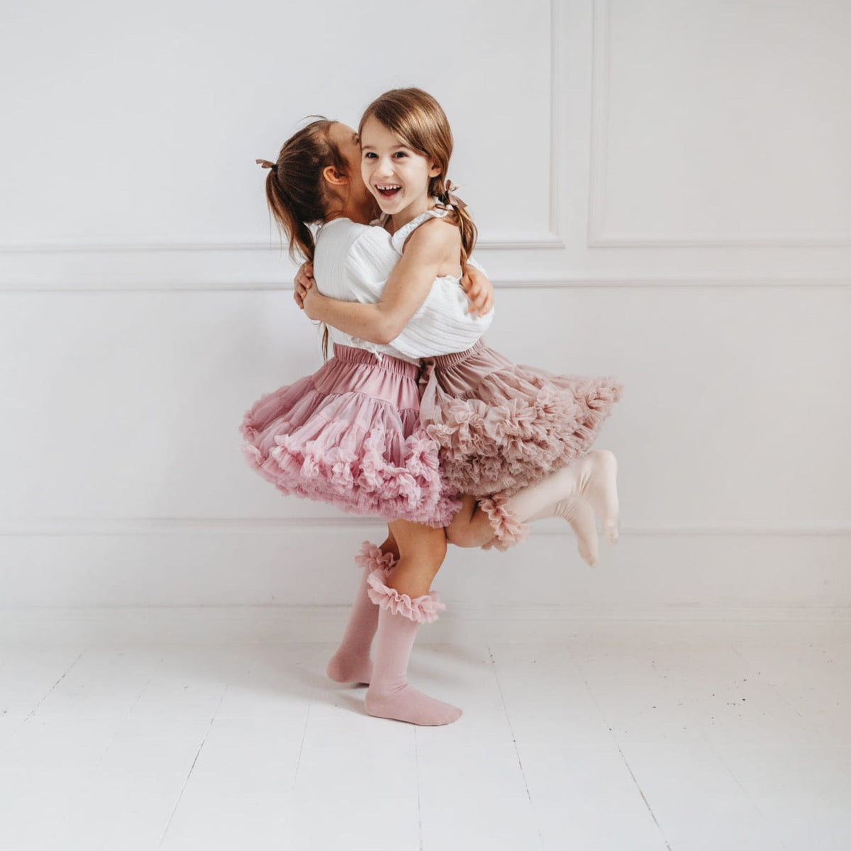 Twee kinderen omhelzen elkaar in tutu rokken en kniekousen met ruches, één in oudroze en één in een lichte tint.