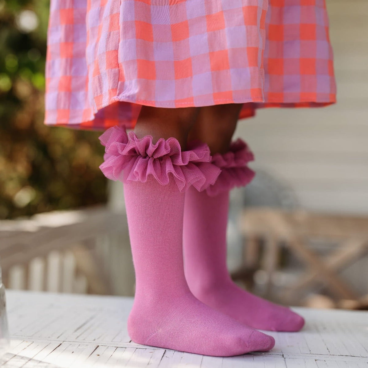 Roze tutu kniekousen met ruches onder een roze geruite jurk, gedragen op een wit houten oppervlak.