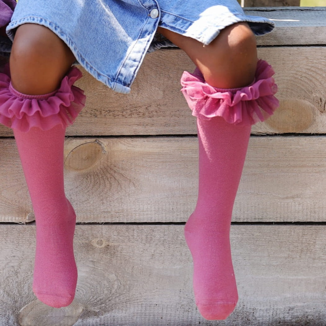 Detail van roze tutu kniekousen met ruches onder een spijkerjurk, met de benen hangend voor een houten wand.