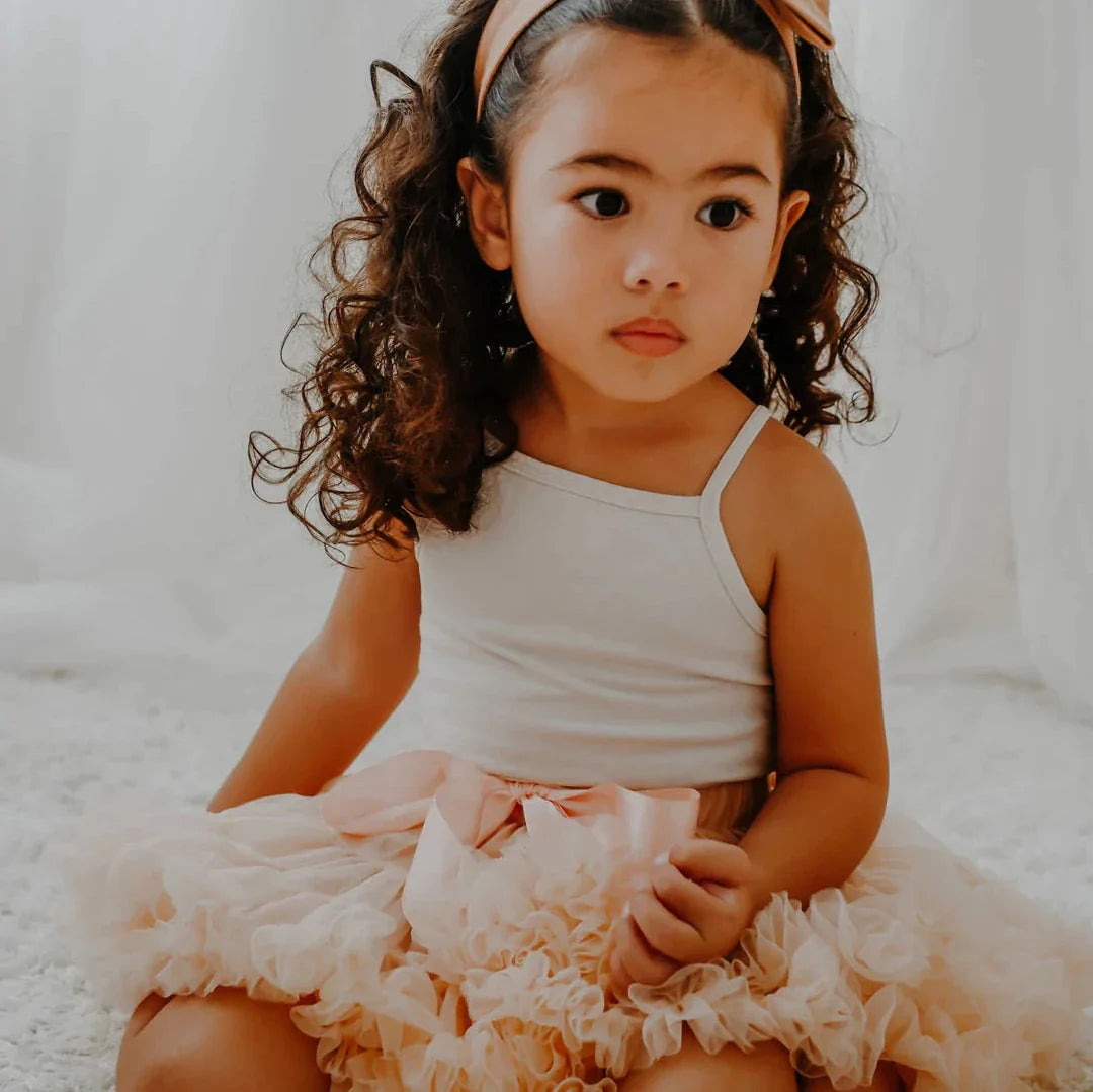 Vooraanzicht van een zittend meisje in de perzikkleurige Loli tutu rok met strik en een witte top.