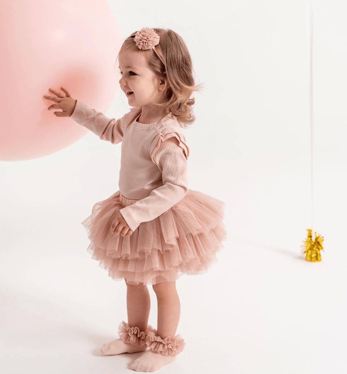 Kind met roze tutu-sokken en een bijpassende rok en haarband, naast een grote roze ballon.