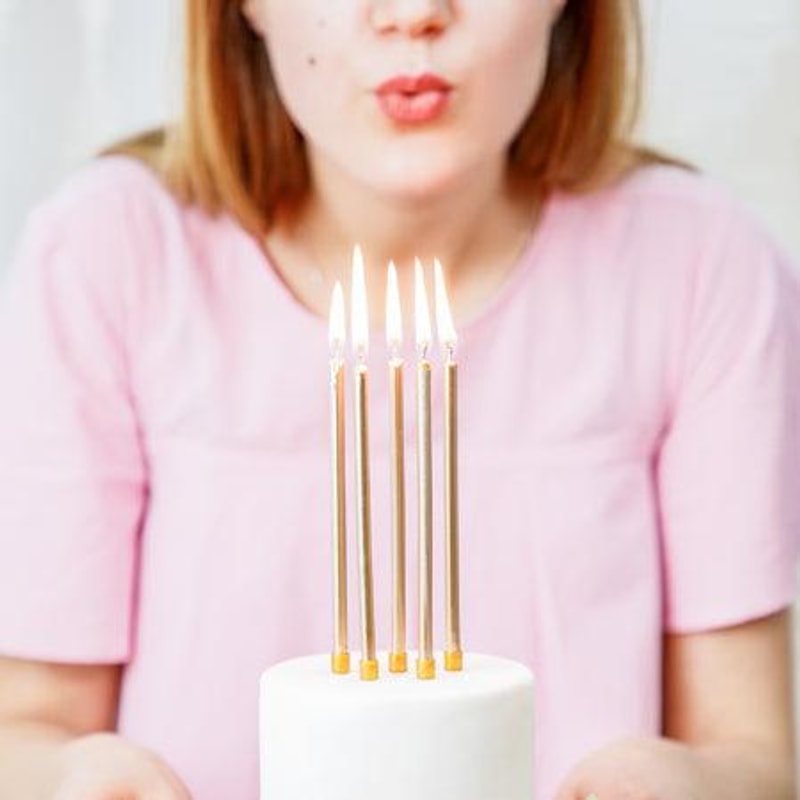 Vrouw blaast naar brandende gouden verjaardagskaarsen op een witte taart.