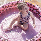 Lachend kind zit in het water van een roze babyzwembad met luipaardprint.
