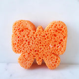 Oranje badspons in vlindervorm met duidelijk zichtbare poriën op een witte ondergrond.