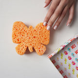 Een hand raakt de oranje badspons in vlindervorm aan, naast een doos met kleurrijke dierenpatronen.