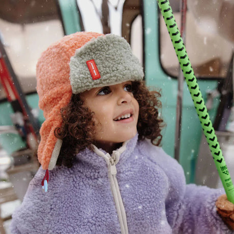 Kind met oranje en grijsgroene Cozy Jim wintermuts en paarse jas, met een groene skistok in de sneeuw.
