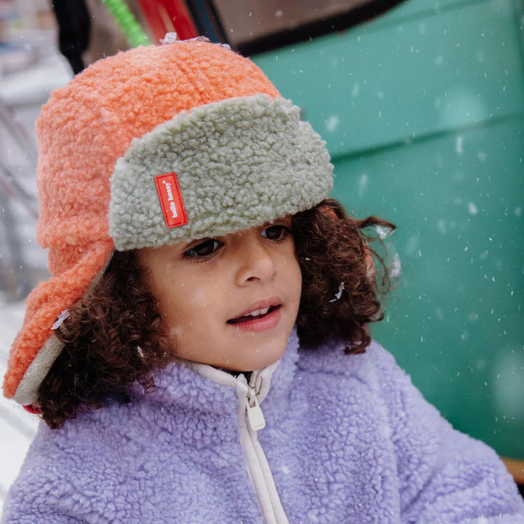 Close-up van een kind met de oranje en grijsgroene Cozy Jim wintermuts met oorkleppen in de sneeuw.