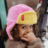 Kind met roze en gele Cozy Kira muts met oorkleppen en een bruine jas in de sneeuw.