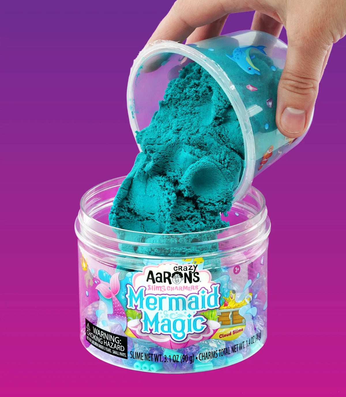 Een hand kantelt een bakje groenblauw Mermaid Magic wolkenslijm boven de open pot.