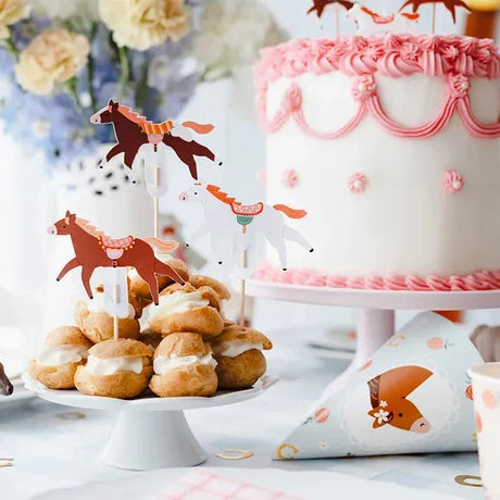 Cupcake toppers met bruine en witte paarden op gebakjes op een witte schaal, naast een taart met roze versiering.