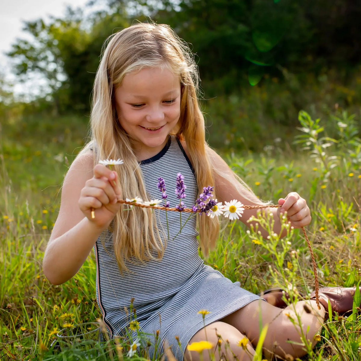 Meisje zit in het gras en versiert een bloemenketting met witte en paarse bloemen.