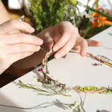 Handen bevestigen bloemen en groene takjes aan een bloemenketting op een witte tafel.