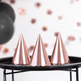 Drie roségouden feesthoedjes op een zwarte tafel met ballonnen en feestdecoratie op de achtergrond