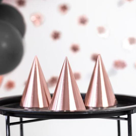 Drie roségouden feesthoedjes op een zwarte tafel met ballonnen en feestdecoratie op de achtergrond