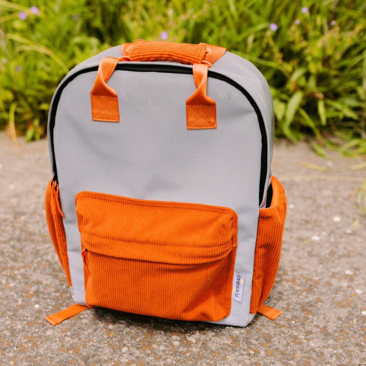 Flashbag Charlie rugzak in grijs met oranje handvatten, voorvak en zijvakken, op een pad in het groen.