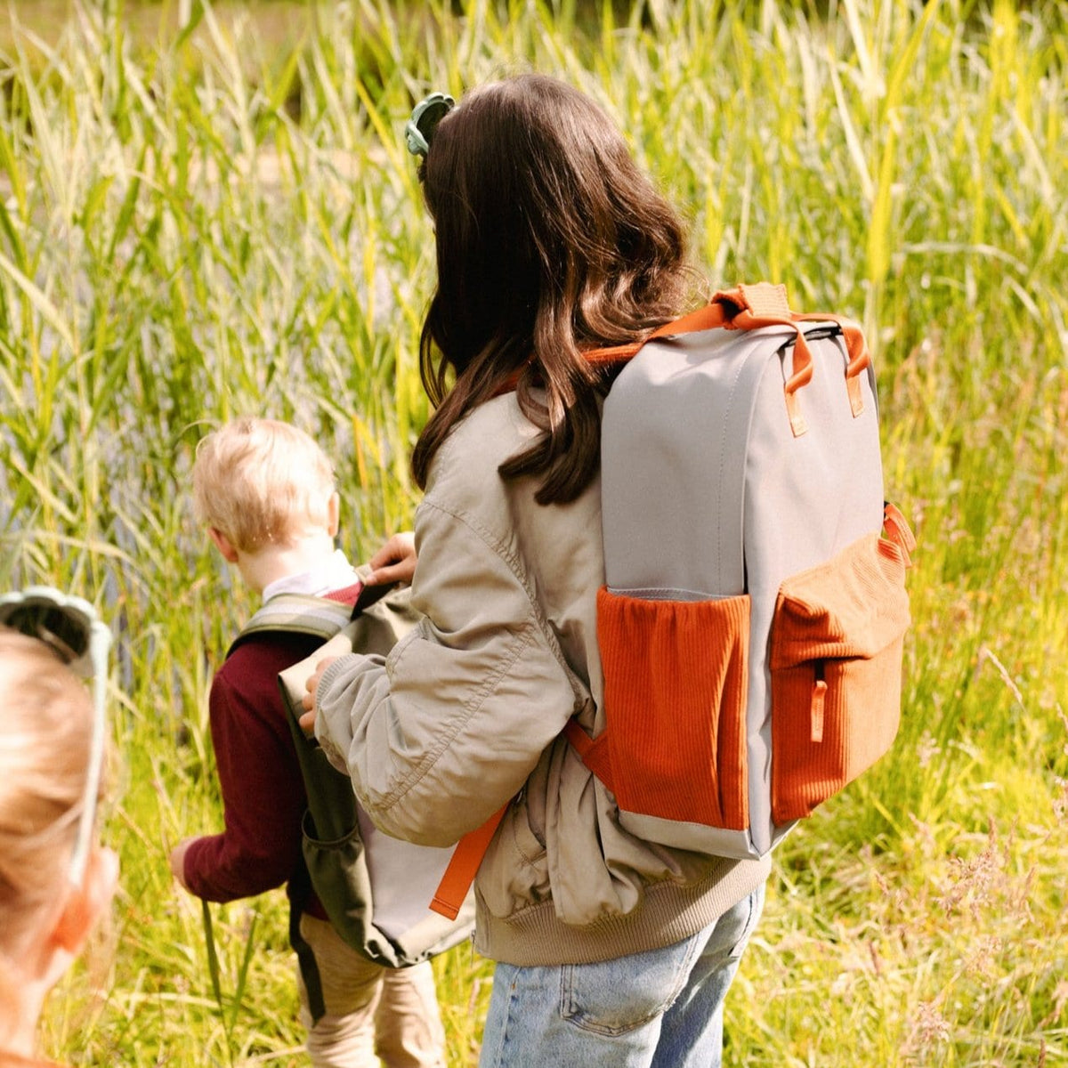 Persoon met grijze Flashbag Charlie rugzak met oranje vakken op de rug, naast een kind tussen het hoge gras.