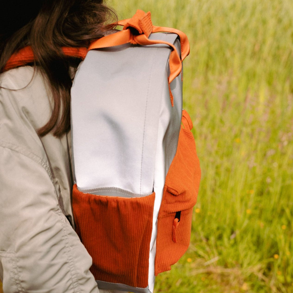 Zijaanzicht van de gedragen Flashbag Charlie rugzak met grijze stof, oranje zijvak en voorvak.