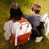 Flashbag Charlie rugzak in grijs en oranje, gedragen door een persoon die naast een kind in het gras hurkt.