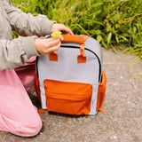 Grijze Flashbag Charlie rugzak met oranje vakken op een pad, naast iemand met een gele bloem in de hand.