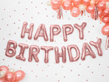Roségouden Happy Birthday-folieballonnen aan een muur met stippen, omringd door roze en witte ballonnen