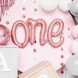 Roségouden folieballon met het woord 'one' tegen een roze achtergrond met sterren en ballonnen.