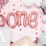 Roségouden folieballon met het woord 'one' tegen een roze achtergrond met sterren en ballonnen.