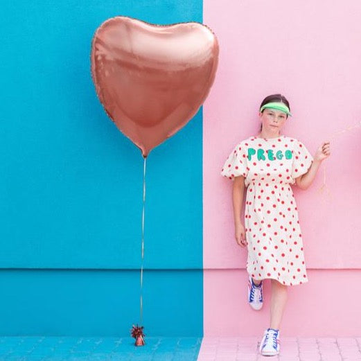 Roségouden hartvormige folieballon aan een lint naast een vrouw, tegen een turquoise en roze muur
