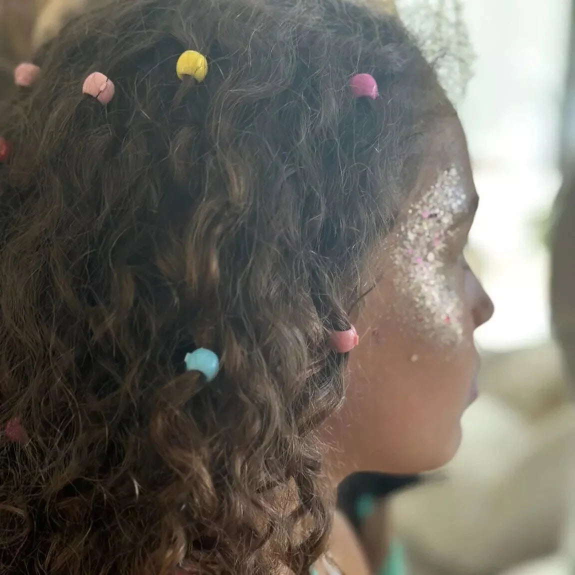 Roze, gele en blauwe haarparels in het krullende haar van een kind met glitter op de wang