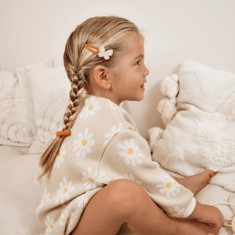 Meisje met een oranje haarspeldje met een beige vlinder naast haar vlecht, zittend op een bank.