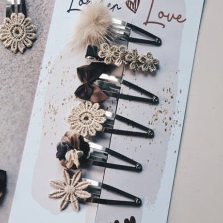 Zes haarspeldjes Stunning Magic op een kaart, versierd met goudkleurige bloemen, bruine strikjes en een pluizig bolletje.