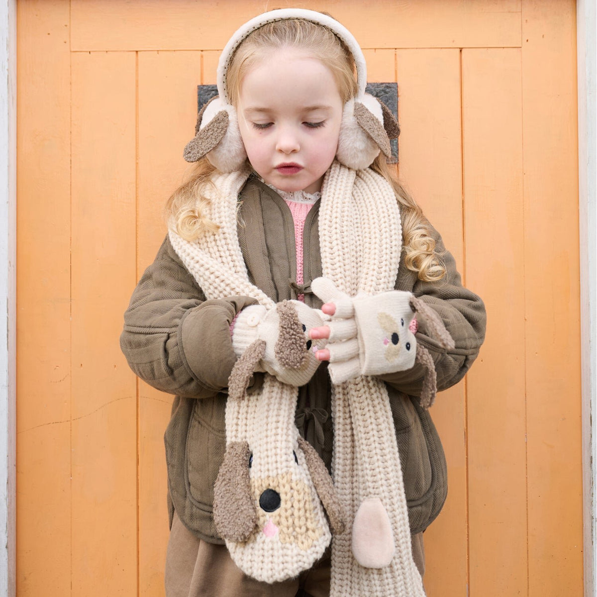 Meisje met beige puppyhandschoenen met open kap, een bijpassende sjaal en oorwarmers voor een oranje deur.